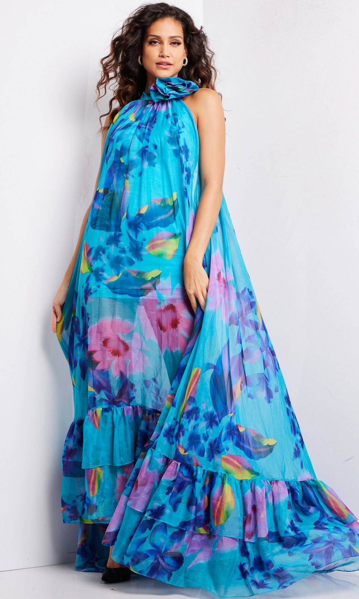 A woman wearing the Long Prom Dress 38721 by Jovani—a flowing blue chiffon formal dress with a floral pattern—stands against a light-colored wall.